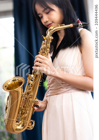 Young woman saxophonist with an instrument stands by the window. Cozy home interior. Concept of hobbies, inner world, development of talents, music school. Young woman saxophonist with an instrument stands by the window. Cozy home interior. Concept of hobbies, inner world, development of talents, music school. 109444496
