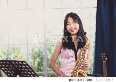 Young woman saxophonist with an instrument stands by the window. Cozy home interior. Concept of hobbies, inner world, development of talents, music school. 109444502