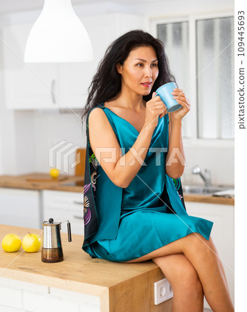 Woman in nightie sitting on table at home and drinking tea Woman in nightie sitting on table at home and drinking tea 109445693