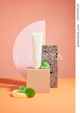 Front view of a white plastic tube unbranded on podiums, green leaves of gotu kola and acrylic sheet on pink background. Mockup for cosmetic of natural extract. Space for design 109449424