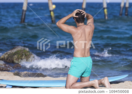Back view of kneeling man on the beach with surfboard resting and looking at the sea before surfing Back view of kneeling man on the beach with surfboard resting and looking at the sea before surfing 109450008