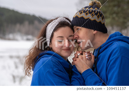 Outdoor happy couple in love posing in cold winter weather. A man and a woman in blue hoodies. Emotional young couple having fun while walking by winter forest, loving man hugging his laughing woman 109451137