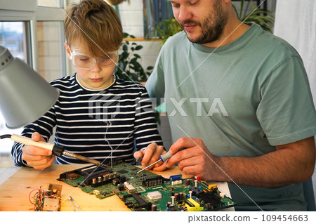 Father with his son spend time together. Close-up of a man teaching his boy at home to solder computer spare parts. Education moment during parenthood. Togetherness concept. 109454663