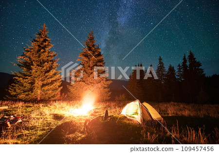Night camping in mountains under starry sky and Milky way. Female tourist resting near burning campfire and illuminated tent in campsite, lying on grass and enjoying night sky with stars. Night camping in mountains under starry sky and Milky way. Female tourist resting near burning campfire and illuminated tent in campsite, lying on grass and enjoying night sky with stars. 109457456