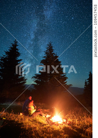 Night camping in mountains under starry sky. Two people, couple sitting on grass near burning campfire, enjoying beautiful sky full of stars. Concept of tourism and healthy living. Night camping in mountains under starry sky. Two people, couple sitting on grass near burning campfire, enjoying beautiful sky full of stars. Concept of tourism and healthy living. 109457458
