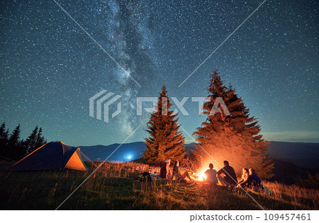Night camping in mountains under starry sky. Back view of group of people traveling. Friends sitting in campsite near fire, talking, enjoying nature. Concept of tourism, adventure and traveling. Night camping in mountains under starry sky. Back view of group of people traveling. Friends sitting in campsite near fire, talking, enjoying nature. Concept of tourism, adventure and traveling. 109457461
