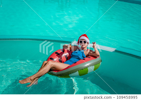 A happy woman in a blue bikini, a red and white Santa hat and sunglasses poses in the pool in an inflatable circle with a watermelon pattern, holding a glass of champagne in her hands. Christmas 109458995