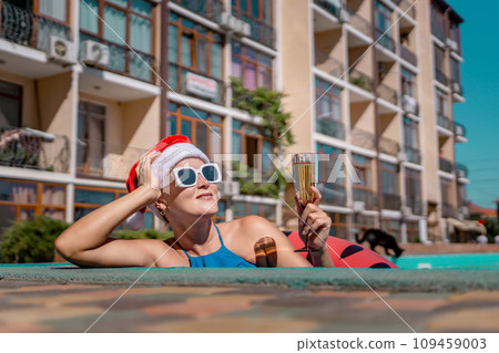 Woman pool Santa hat. A happy woman in a blue bikini, a red and white Santa hat and sunglasses poses near the pool with a glass of champagne standing nearby. Christmas holidays concept. Woman pool Santa hat. A happy woman in a blue bikini, a red and white Santa hat and sunglasses poses near the pool with a glass of champagne standing nearby. Christmas holidays concept. 109459003