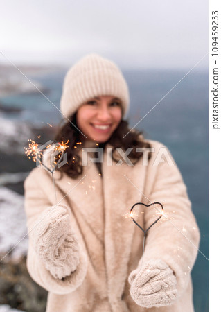 Outdoor winter portrait of happy smiling woman, light faux fur coat holding heart sparkler, posing against sea and snow background Outdoor winter portrait of happy smiling woman, light faux fur coat holding heart sparkler, posing against sea and snow background 109459233