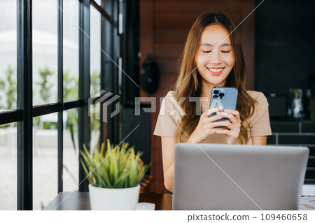 Happy lifestyle female smiling using smartphone in coffee shop, young business woman working with laptop computer she holding smart mobile phone at cefa for texting messages Happy lifestyle female smiling using smartphone in coffee shop, young business woman working with laptop computer she holding smart mobile phone at cefa for texting messages 109460658