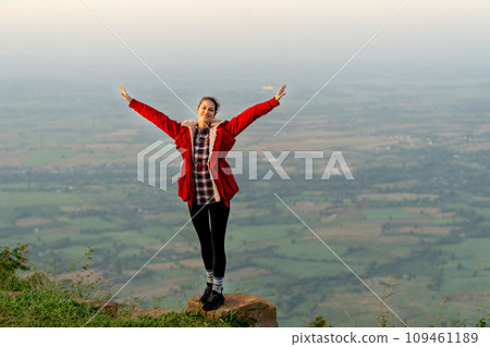 Wide shot of woman wear red coat and stand with spread her arms near cliff on the mountain and look at camera with beautiful high view background with warm light of sun rising. Wide shot of woman wear red coat and stand with spread her arms near cliff on the mountain and look at camera with beautiful high view background with warm light of sun rising. 109461189