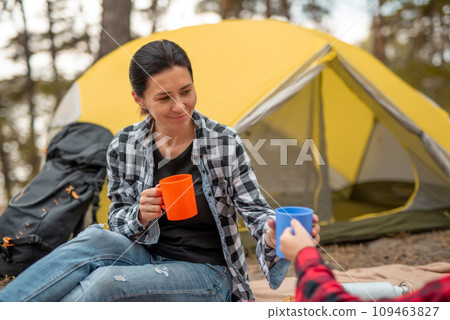 Beautiful adult girls drinking tea in camping in forest with tent on background and smiling. Young woman traveller during hiking in mountains resting 109463827