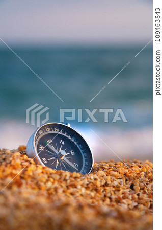 Conceptual photo, close-up of a compass lying in wet sand consisting of small crumbs of shells, against the backdrop of a blue calm sea Conceptual photo, close-up of a compass lying in wet sand consisting of small crumbs of shells, against the backdrop of a blue calm sea 109463843
