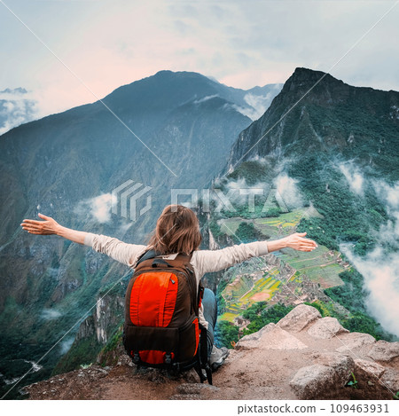 View of girl sitting on the edge of the rock with raised hands and taking pleasure of breathtaking forested mountains landscape of Machu Picchu View of girl sitting on the edge of the rock with raised hands and taking pleasure of breathtaking forested mountains landscape of Machu Picchu 109463931