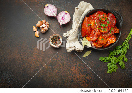 Cooking hearty meat stew. Raw uncooked chopped pieces of meat marinated with seasonings and parsley in black casserole dish top view, ingredients on dark rustic background. Space for text Cooking hearty meat stew. Raw uncooked chopped pieces of meat marinated with seasonings and parsley in black casserole dish top view, ingredients on dark rustic background. Space for text 109464181
