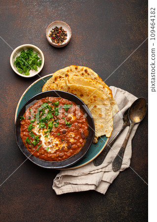 Traditional Indian Punjabi dish Dal makhani with lentils and beans in black bowl served with naan flat bread, fresh cilantro and two spoons on brown concrete rustic table top view 109464182