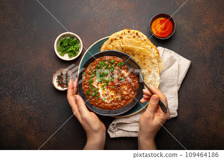 Female hands holding a bowl and eating traditional Indian Punjabi dish Dal makhani with lentils and beans served with naan flat bread, fresh cilantro on brown concrete rustic table top view 109464186