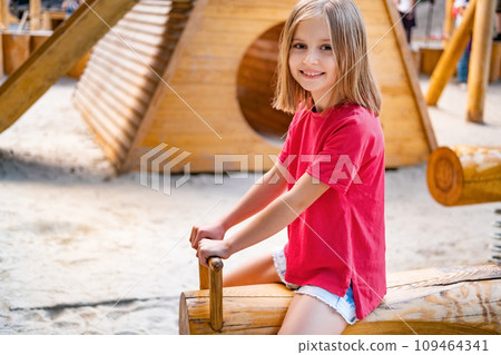 Happy little girl riding wooden swing on kids playground Happy little girl riding wooden swing on kids playground 109464341