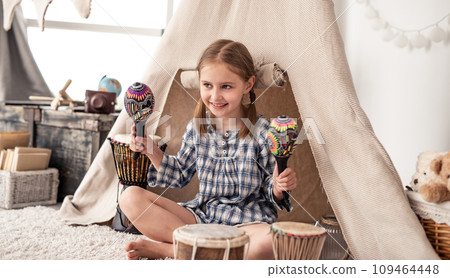Happy little girl playing on wooden maracas in kids room decorated with ethnic drums and wigwam Happy little girl playing on wooden maracas in kids room decorated with ethnic drums and wigwam 109464448