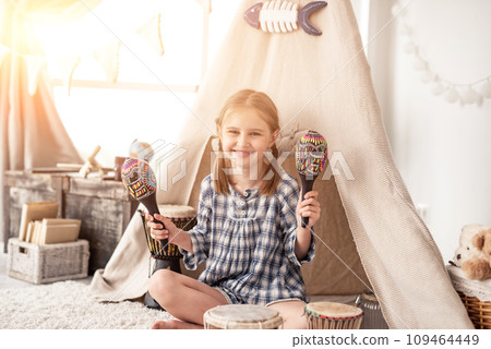 Happy little girl playing on wooden maracas in room decorated with ethnic drums and wigwam 109464449