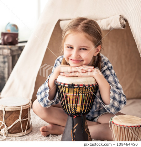 Portrait of Little girl playing on traditional african djembe drums sitting in wigwam in kids room Portrait of Little girl playing on traditional african djembe drums sitting in wigwam in kids room 109464450