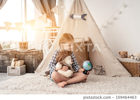 Little girl exploring globe with teddy bear sitting in wigwam in playroom 109464463