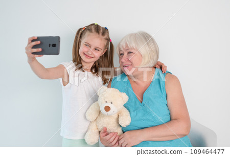 Smiling little girl taking selfie with grandmother against white wall at home Smiling little girl taking selfie with grandmother against white wall at home 109464477