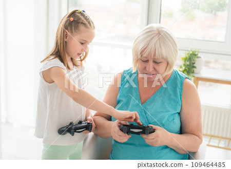 Little girl teaching elderly woman to play on joystick indoors Little girl teaching elderly woman to play on joystick indoors 109464485