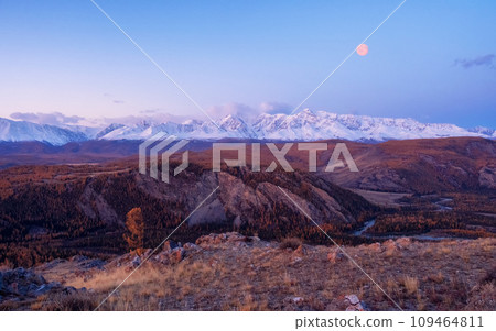 Dawn in the mountains with a full moon in the sky. Impressive panorama of dawn snow-capped peaks and a wide mountain valley with winding river in a canyon in soft purple tones. Dawn in the mountains with a full moon in the sky. Impressive panorama of dawn snow-capped peaks and a wide mountain valley with winding river in a canyon in soft purple tones. 109464811