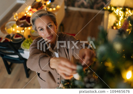 Top view of elegant middle-aged woman hangs up ball on branch of xmas tree in cozy living room, looking at camera. Happy female house decoration and preparing to celebrate Christmas and New Year. 109465676