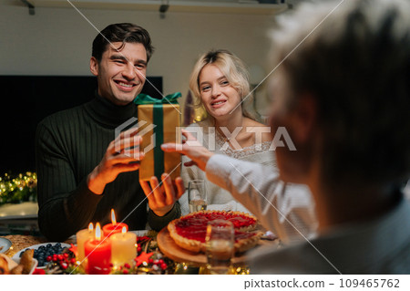 View from back of unrecognizable mother-in-law giving festive gift box with Christmas present to happy man sitting with family at dinner table at home on xmas eve. Concept of home festive atmosphere. View from back of unrecognizable mother-in-law giving festive gift box with Christmas present to happy man sitting with family at dinner table at home on xmas eve. Concept of home festive atmosphere. 109465762