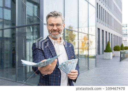 Satisfied businessman holding a lot of money in his hands, stretching it forward, winning the lottery, investment, smiling and looking at the camera. 109467470