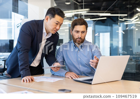 Two businessmen in suits work in the office, look at a laptop, discuss work issues, have a discussion. Two businessmen in suits work in the office, look at a laptop, discuss work issues, have a discussion. 109467519