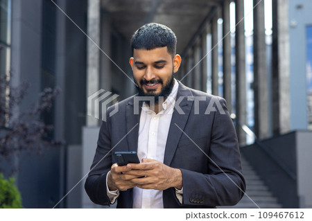 Indian young male businessman standing on the street near an office building and using a mobile phone, dialing a number, writing a message, reading the news. 109467612