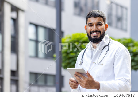Portrait of a young smiling male Muslim doctor standing in a white coat and with a stethoscope outside a clinic and using a tablet. Portrait of a young smiling male Muslim doctor standing in a white coat and with a stethoscope outside a clinic and using a tablet. 109467618
