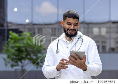 Smiling young male arab doctor standing outside on the street near the clinic and using a tablet. Smiling young male arab doctor standing outside on the street near the clinic and using a tablet. 109467623