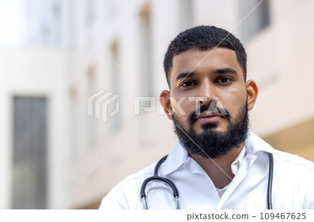 Close-up portrait of a young Muslim male doctor standing outside a clinical center in a white coat and looking seriously into the camera. Close-up portrait of a young Muslim male doctor standing outside a clinical center in a white coat and looking seriously into the camera. 109467625