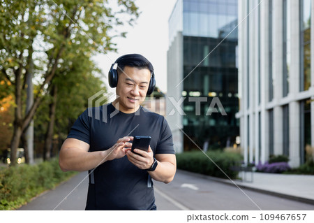 A young Asian man in sportswear is standing on a city street wearing headphones and using a mobile phone. A young Asian man in sportswear is standing on a city street wearing headphones and using a mobile phone. 109467657