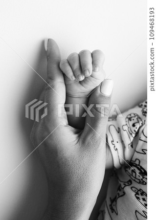 Parents' hands hold the fingers of a newborn baby. The hand of a mother and father close-up holds the fist of a newborn baby. Family health and medical care. Tiny fingers in a black and white photo. 109468193