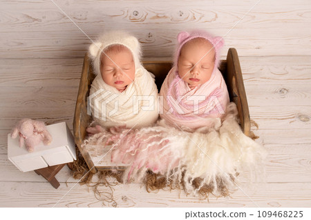Tiny newborn twin girls in a pink and white cocoon in a cute wooden crib against the background of old vintage white boards. Newborn twin girls with white and pink hats with ears on their heads.  109468225