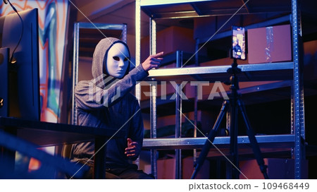 Hacker wearing anonymous mask doing ransom video after gathering information from users computers using spyware software. Cybercriminal blackmailing victim in graffiti painted hideout Hacker wearing anonymous mask doing ransom video after gathering information from users computers using spyware software. Cybercriminal blackmailing victim in graffiti painted hideout 109468449