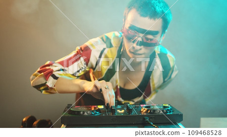 Young cool artist working as dj with turntables, mixing modern remix with bass and audio stereo equipment. DJ performer playing music with mixer and disc buttons. Handheld shot. Young cool artist working as dj with turntables, mixing modern remix with bass and audio stereo equipment. DJ performer playing music with mixer and disc buttons. Handheld shot. 109468528