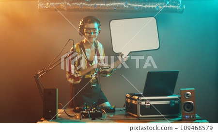 Woman mixer holds speech bubble icon at nightclub, performing techno music mix on stage. Artist using turntables to create mixing show, presenting isolated cardboard placard. Tripod shot. 109468579
