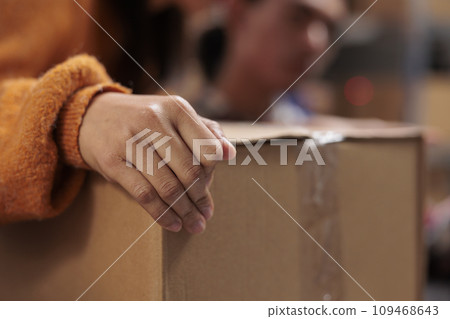 Package handler arm holding packed cardboard box in industrial warehouse. Storehouse employee picking order and preparing customer parcel for transportation with close up on worker hand 109468643