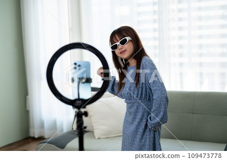 A cheerful young Asian girl in a cute dress and sunnies is recording her dance video at home. A cheerful young Asian girl in a cute dress and sunnies is recording her dance video at home. 109473778