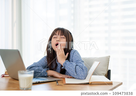 A cute young Asian girl is feeling tired and sleepy, yawning while studying online in her room. 109473838