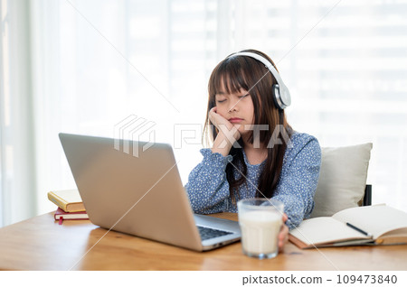 A cute young Asian girl is falling asleep while studying online at a table in her room. A cute young Asian girl is falling asleep while studying online at a table in her room. 109473840