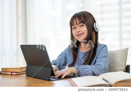 An adorable young Asian girl is studying online on her digital tablet at a table in her room. An adorable young Asian girl is studying online on her digital tablet at a table in her room. 109473841
