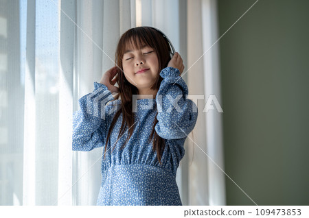 A cute and happy young Asian girl is enjoying listening to music on headphones in the living room. 109473853