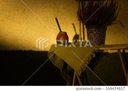 Mate calabash with bombilla. Healthy infused drink, classic Yerba mate tea on a chair on a cozy patio, backyard. Hot beverage on a terrace. Traditional Latin America cuisine. Useful drink still life. 109474817